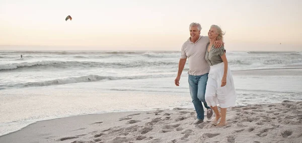 Mature married couple walking on the beach. Senior man hugging his wife by the ocean. Mature couple strolling on the beach at sunset. Older couple being affectionate on holiday by the ocean.
