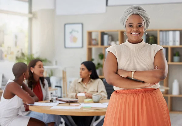 Portrait of smiling african american plus size business woman standing with her arms folded while colleagues sit behind her in office. Ambitious, confident, happy black professional with arms crossed.