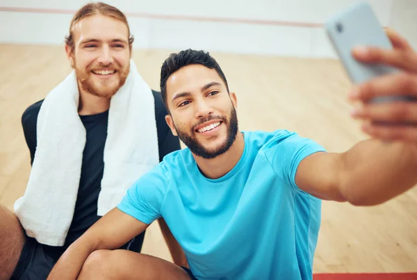 Two young athletic squash players using cellphone for selfie after playing court game. Smiling fit active Caucasian and mixed race athlete sitting together and feeling happy while posing for picture.