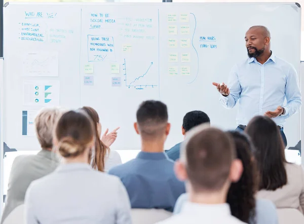 Mature african american business manager training and teaching team of colleagues in office. Black businessman using a whiteboard to talk and explain to diverse group of business people in workshop.