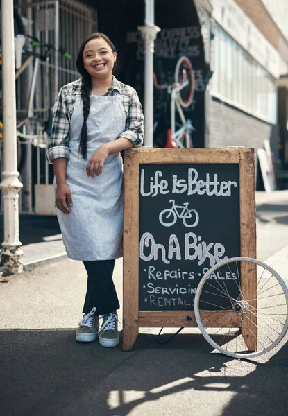 Everyone finds their place eventually. Portrait of a young woman leaning on a bicycle repair shop sign outside