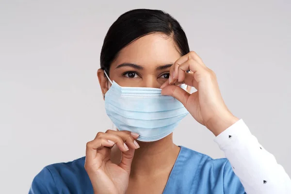 Love for medicine equates love for humanity. Portrait of a young doctor adjusting her surgical face mask against a white background