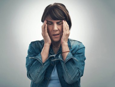 Üzgünüm, migrenim var. Geçici olarak düzensizim. Yaşlı bir kadının gri bir arka planda baş ağrısı çekerken çekilmiş bir fotoğrafı.