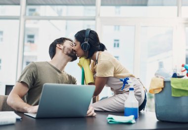 Serbest çalışmanın neden harika olduğunu bilmek isteyen herkes için. Genç bir kadın evi temizlerken kocasını öpüyor ve kocası dizüstü bilgisayar kullanıyor.