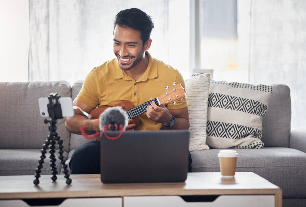 Guitar, streaming and phone with a man talking online to coach during live lesson. Asian male influencer happy on home sofa with a ukulele as content creator teaching music on education podcast blog.