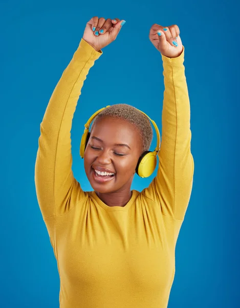 Black woman with headphones, dance and listening to music with rhythm and fun with freedom on blue background. Happy female with arms raised, streaming radio with dancing and carefree in studio.