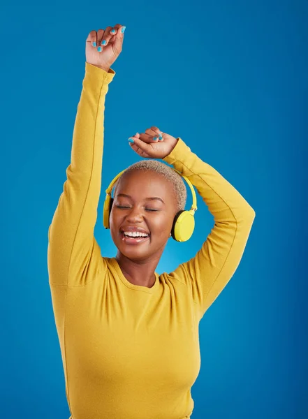 Black woman with headphones, dance and listen to music with rhythm and fun with freedom on blue background. Happy female with arms raised, streaming radio with dancing and carefree in studio.