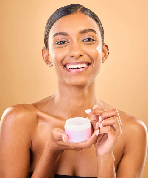 Face, skincare and woman with cream jar in studio isolated on a brown background. Dermatology cosmetics, portrait and happy Indian female apply lotion, creme and moisturizer product for healthy skin