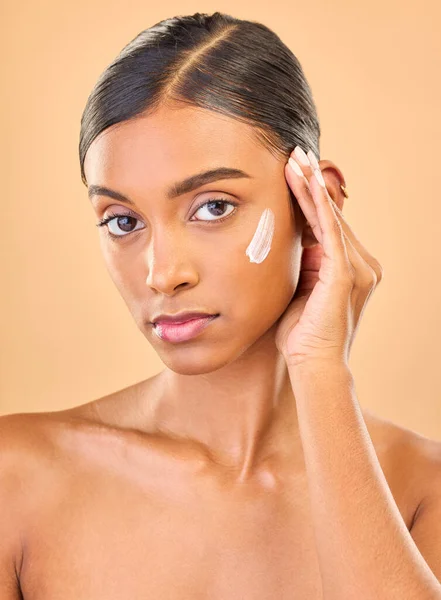 stock image Face portrait, skincare and woman with cream in studio isolated on brown background. Dermatology, serious cosmetics and confident Indian female model with lotion, creme or moisturizer for skin health.