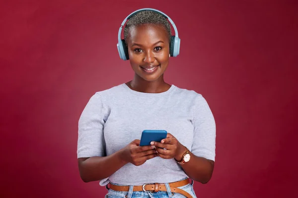 Happy black woman, phone or headphones for music, podcast or listening to radio in red studio background. Smile, bluetooth or wireless girl for relax audio, 5g smartphone or social media networking.
