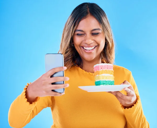 Woman, cake and smile selfie in studio for social media profile picture and excited to eat. Happy female laughing on blue background with rainbow color dessert for birthday or influencer celebration.