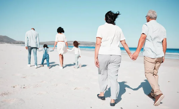 Senior couple, holding hands and family at beach with back for walk, freedom and vacation together with love. Old man, woman and grandchildren by ocean for walking, wellness and adventure in sunshine.