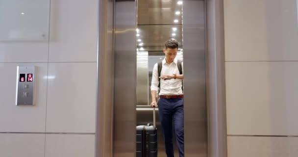 エレベーター 空港でビジネスアジアの男性と一緒に見て 出発のための彼のフライトの時間をチェックしてください 旅行中にスーツケースの荷物を保持男性従業員と後半 — ストック動画