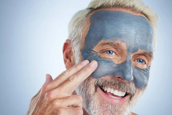 Stock image Senior man, skin and face mask in studio for health, wellness and organic cleaning with smile by background. Elderly male, hand and natural charcoal product for facial cosmetics for dermatology detox.