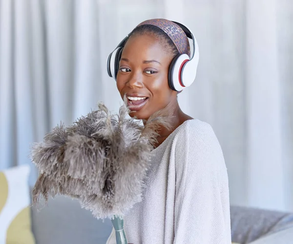 Happy, black woman and cleaning with headphones music, singing and enjoying radio while housekeeping. Smile, duster and an African girl listening to a podcast or audio to clean the living room.