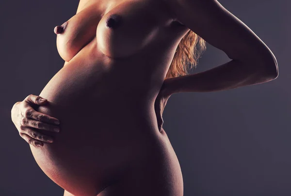 Pregnancy, naked woman in studio with hand on stomach and aesthetic dark background at maternity reveal. Creative photoshoot, art and nude pregnant mother holding belly with healthy body and wellness.