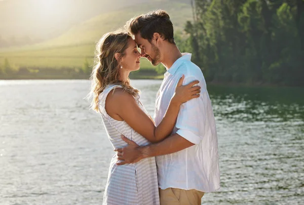 Couple, head touch and hug by lake for outdoor date, romantic adventure and summer love in nature. Man, woman and romance with embrace, care and bond by water with sunshine, freedom and happiness.