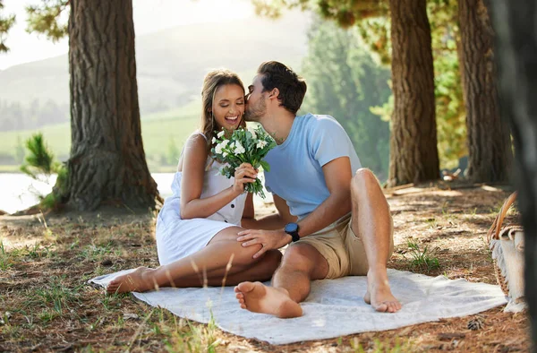 Couple kiss, love and flowers on picnic, summer with freedom and adventure, affection in relationship and care outdoor. Happy people together in nature, commitment and trust with romance in forest.