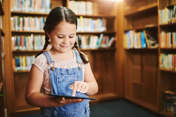 彼女は装置に十分注意を払う 昼間図書館の中に立っている間にタブレットを見ている陽気な若い女の子 — ストック写真