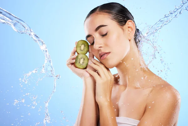 Woman, water splash and face with kiwi in relax for skincare, hygiene or nutrition against blue studio background. Calm or relaxed female holding fruit in natural beauty cosmetics or facial treatment.