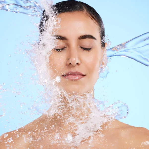 Water splash, face and skincare with a woman in studio on a blue background for hygiene or hydration. Relax, beauty and cleaning with an attractive young female in the shower for self care.