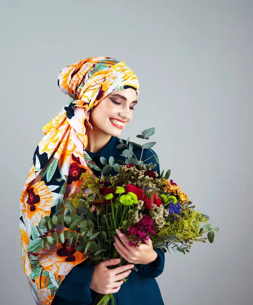 她每天收到这么多的花 一个自信的年轻女子头戴五颜六色的头巾 拿着一束灰色背景的花的摄影棚照片 — 图库照片
