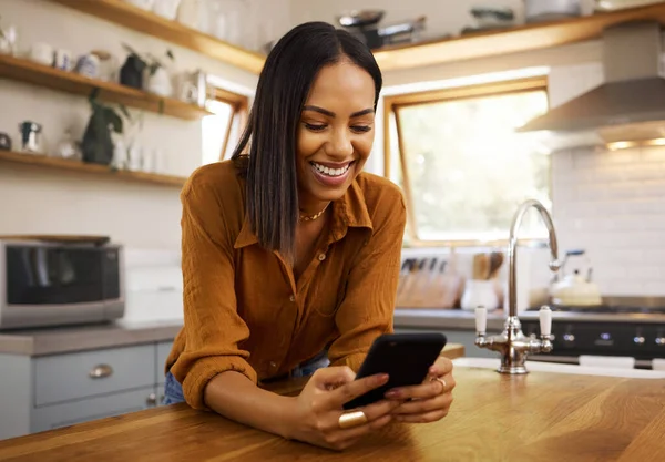 Happy woman, phone text and kitchen in a home reading a web app. House, female and smile of a young person with joy resting on a counter top table feeling relax typing with mobile networking.