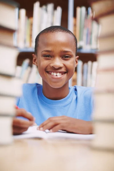 その広い笑顔は彼が学びたいとあなたに伝えます アフリカ系アメリカ人の少年が本の山の間の図書館であなたを笑っています — ストック写真