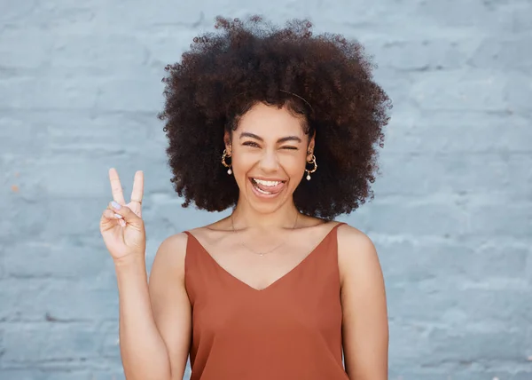 Black woman, portrait and tongue out with peace sign and afro against a gray wall background. Happy and goofy African female face smile showing peaceful hand emoji, sign and funny or silly expression.