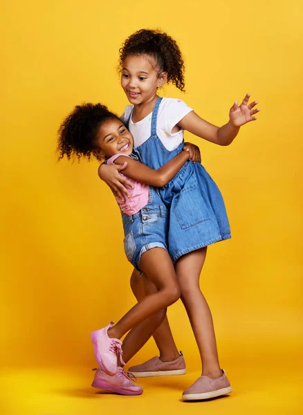 Children, hug and happy sisters in studio dancing with love, care and support of family on yellow background. Cute young girl kids play together for happiness, fun and positive attitude or fashion.