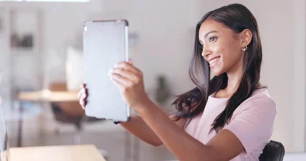 Young happy woman taking a selfie with a digital tablet while relaxing in an office at work. One smiling female student taking pictures to post on social media while sitting alone in a library.