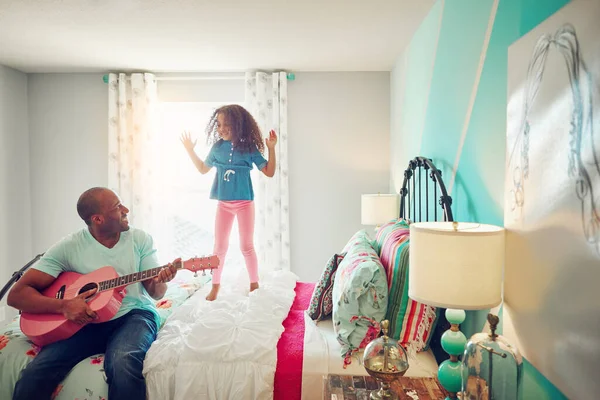 Shes his biggest fan. a cheerful little girl jumping on her bed at home while her father plays the guitar