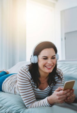 Doğru müzik onu rahatlama moduna sokuyor. Kanepede dinlenen ve telefon ve kulaklıklarını kullanan genç bir kadın.