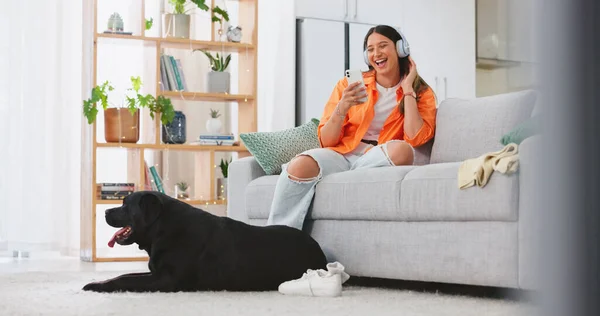 Phone, headphones and laughing woman with dog on sofa in home living room. Comic, relax and happy female streaming music, radio or podcast with smartphone while playing with animal, pet or bonding