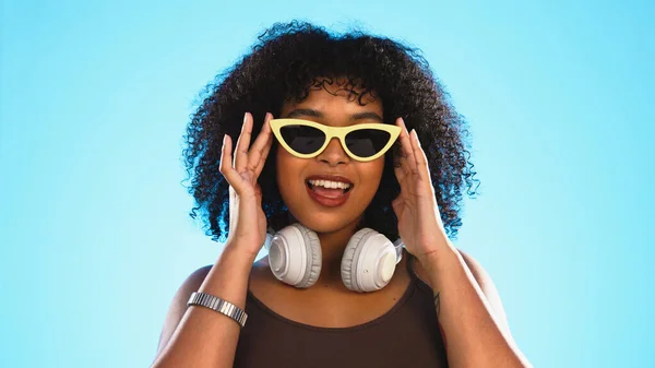 Trendy black woman face isolated on blue background for beauty, cosmetics and fashion of gen z lifestyle. Confident african american person or model with cool sunglasses and headphones for summer.
