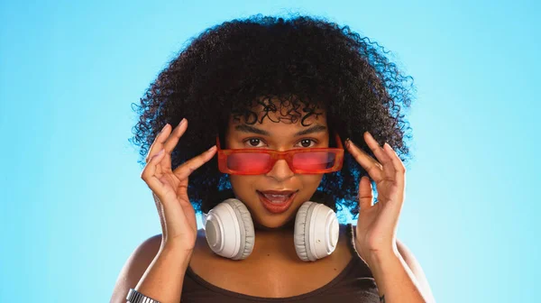 Trendy, gen z and face of a black woman in a studio with accessories, sunglasses and fashion. Happy, afro and portrait of African female model with a cool, stylish and edgy style by a blue background.