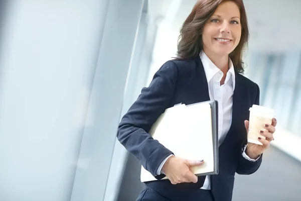 Cheerful businesswoman holding folders and drinking a coffee at work. One female businessperson carrying files and drinking tea on a break in an office. Business professional at a startup company.