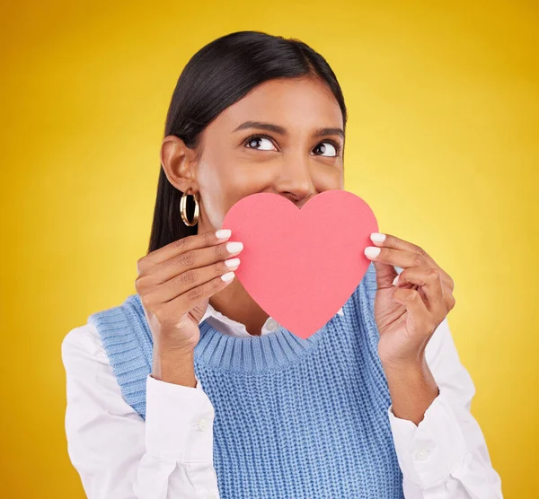 Shy, paper and heart with woman in studio for love, support and romance. Valentines day, kindness and date with female and hiding with symbol on yellow background for health, happiness or hope mockup.