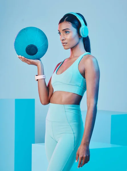 Fitness, ball and woman in studio with music for training, exercise and sports routine on blue background. Headphones, radio and Indian female athlete with weight and podcast, mindset and focus.