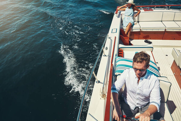 Bon Voyage. High angle shot of a handsome young man steering a yacht with his wife sitting in the background