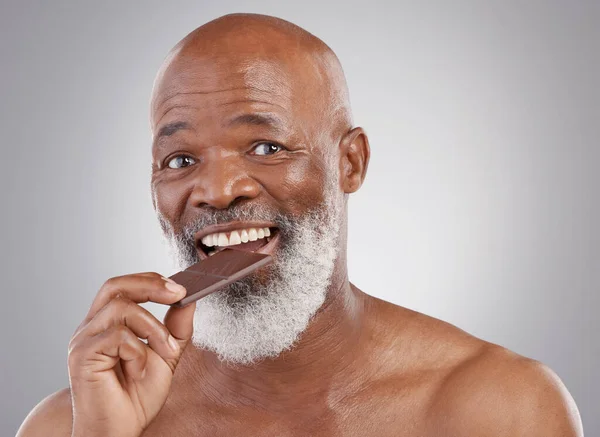 Black man, portrait and eating chocolate sweets isolated on a studio background for a treat. Happy, snack smile and an elderly African model biting into a sweet candy bar for happiness and sugar.