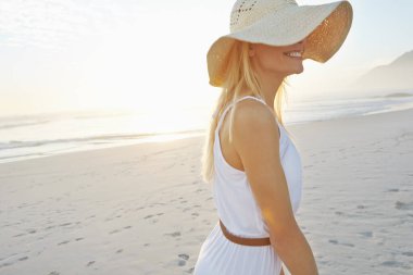 Sahildeki zamanının tadını çıkarıyor. Sahilde beyaz bir elbise ve güneş şapkası takan muhteşem bir genç kadın.