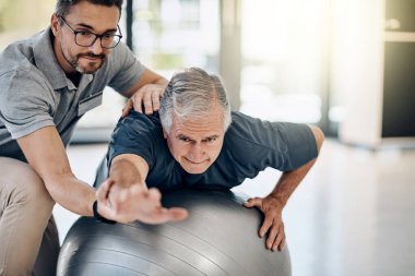 Çekirdek kaslarını hedef alalım. İlgili bir fizyoterapist, olgun hastasına rehabilitasyon merkezinde yoga topu kullanmasında yardım ediyor.