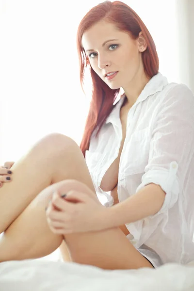 一个漂亮的红头发女人穿着一件未扣子的衬衫坐在床上 看起来很性感 同时摸着她光滑的 光滑的剃光的腿的画像 年轻女子半裸姿势 胸部以下露出 — 图库照片