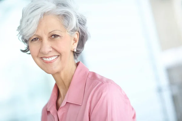 stock image Portrait of senior woman smiling with healthy white teeth. Beautiful old female standing alone inside a room. Happy attractive mature lady with short hair. Pretty older person with good dental care.