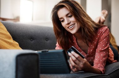 En sevdiğin dükkanlara göz atmak daha da zekileşti. Evdeki kanepede dijital tablet ve kredi kartı kullanan çekici bir genç kadın.