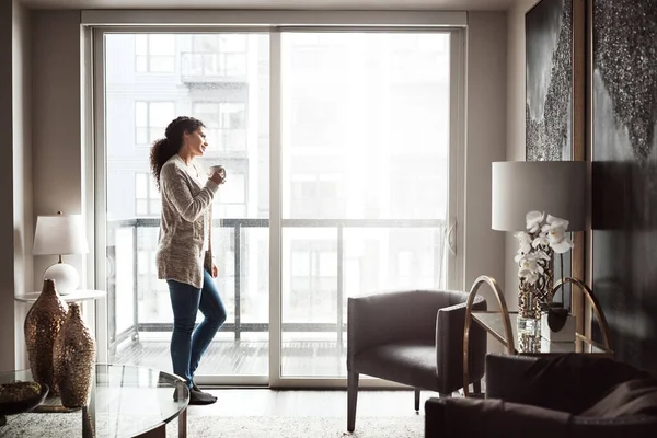 stock image Living alone has its perks. a cheerful young woman drinking coffee while looking through a window inside at home during the day