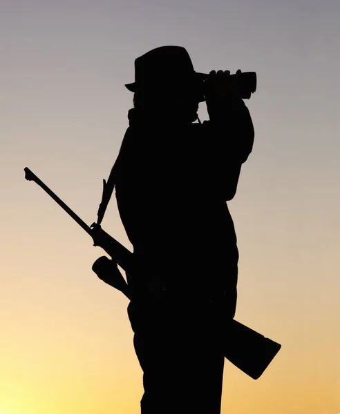 Hunting at sunset, man with gun and binoculars in nature to hunt game for sport on safari adventure. Sky, silhouette and hunter with rifle, search and setting sun for shooting hobby on summer evening.