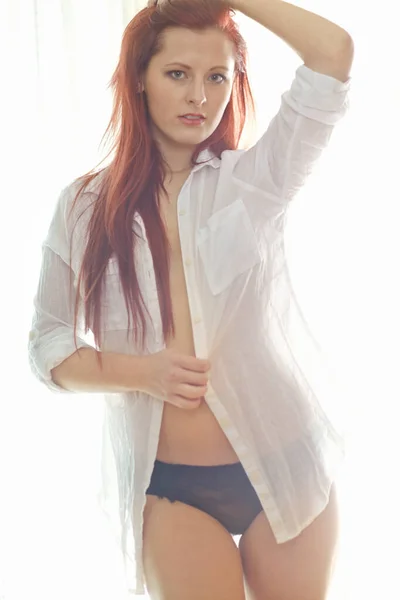 一个漂亮的红头发女人穿着没有扣子的衬衫站在卧室里 站着时看起来很性感的画像和她的内裤 年轻女子半裸着身子 双手高举头顶 — 图库照片