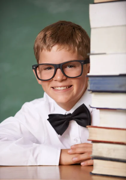 丰富了他年轻的心灵 一个戴着眼镜和领结的小男孩从书堆后面对着摄像机微笑 — 图库照片